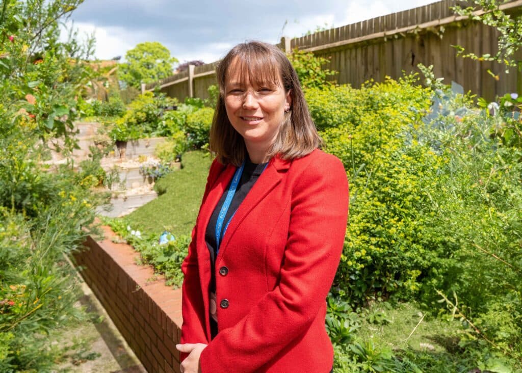 A woman in a red blazer stands outdoors in a garden with green bushes, plants, and a wooden fence in the background, smiling at the camera outside the Nugent Boarding House.