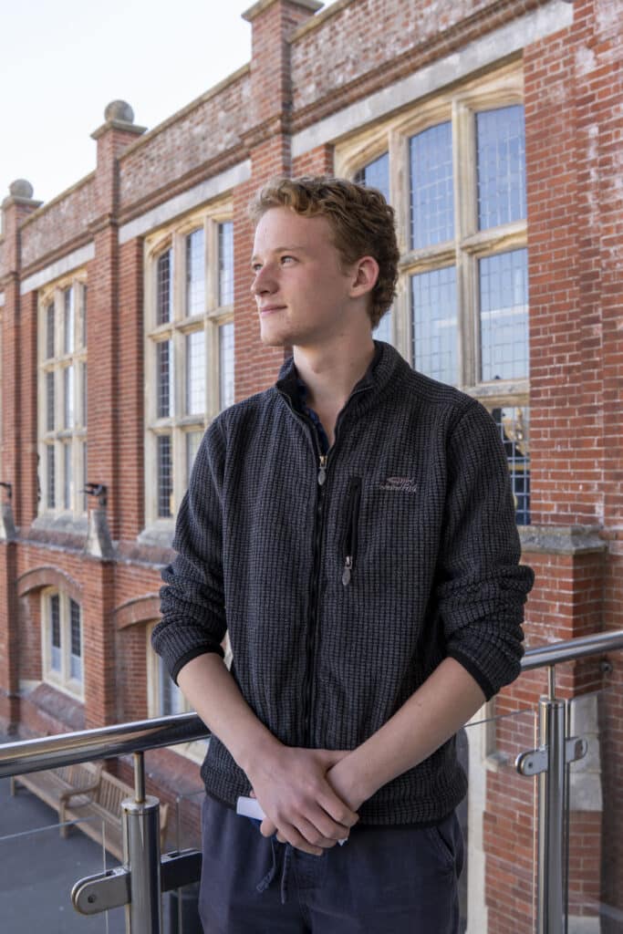 A young man with curly hair stands on a balcony, wearing a dark zip-up jumper, looking off to the side, with a brick building and large windows in the background.