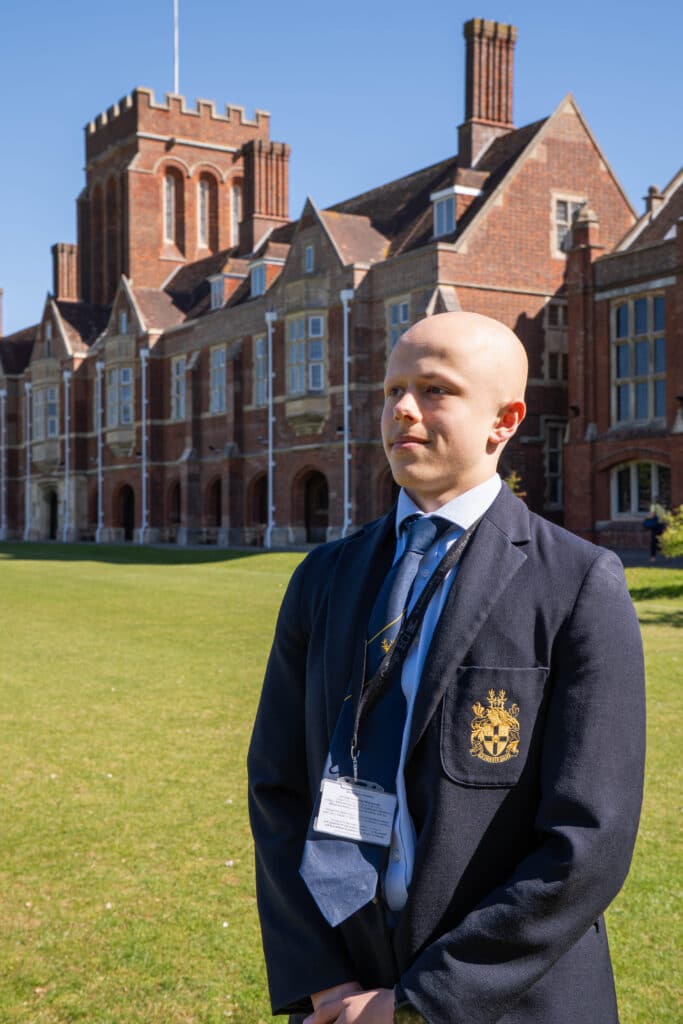 A person in a dark blazer with a crest stands on grass in front of a large brick building with tall windows and chimneys, under a clear blue sky.