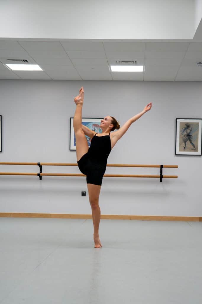 A dancer in a black outfit balances on one foot in a studio, holding her other leg high and straight beside her head, arms extended gracefully. Ballet barres and framed art are visible on the wall behind her.