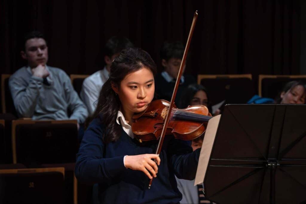 A young woman plays the violin intently on stage, reading sheet music. Several people sit in the darkened audience behind her, watching quietly.