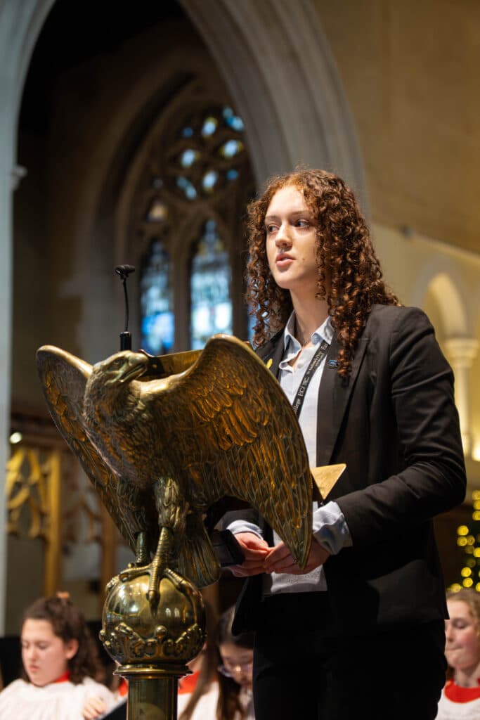 Humreen Ellens, a person with curly hair, stands at a lectern shaped like an eagle, speaking in a church. Stained glass windows and seated people in white robes are visible in the background.