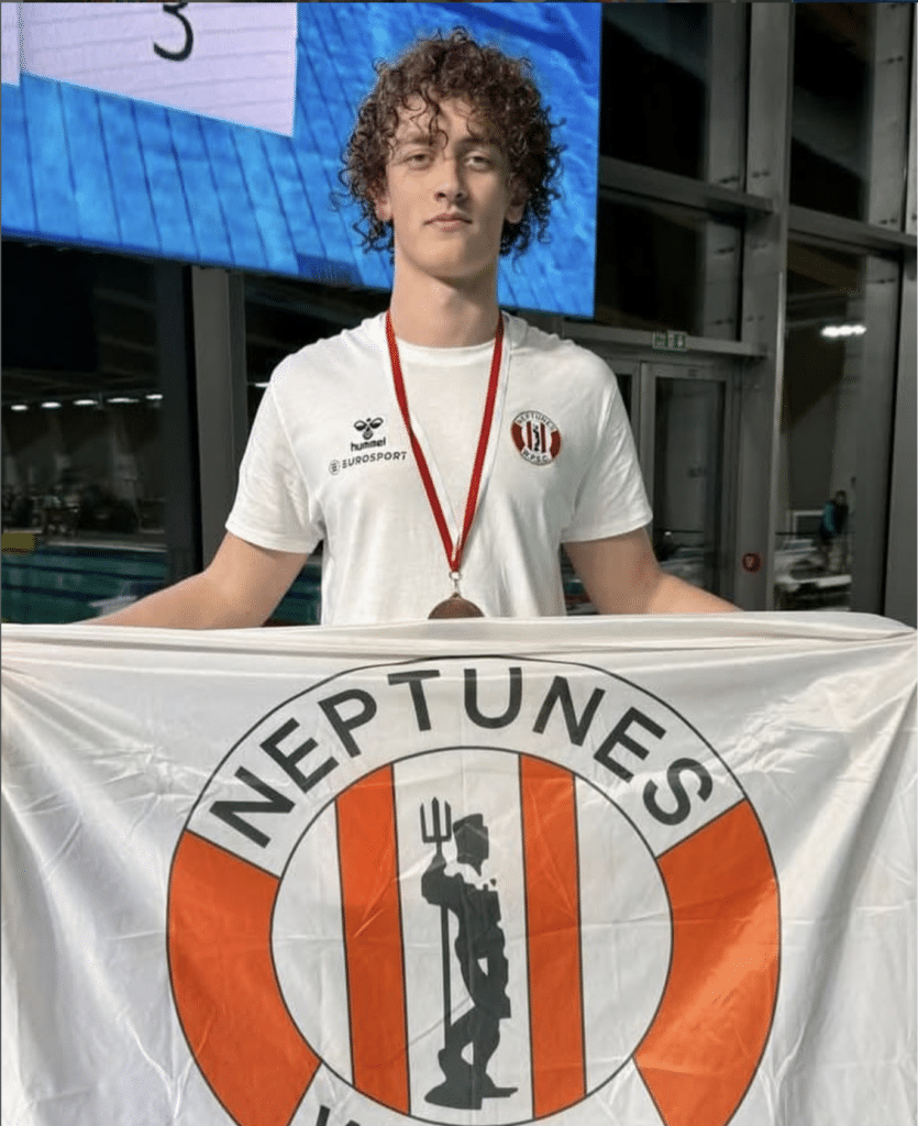 Young athlete Jack Hylands with curly hair holds a Neptunes flag and wears a medal round his neck. He stands indoors by a pool, wearing a white shirt with a sports logo and badge.