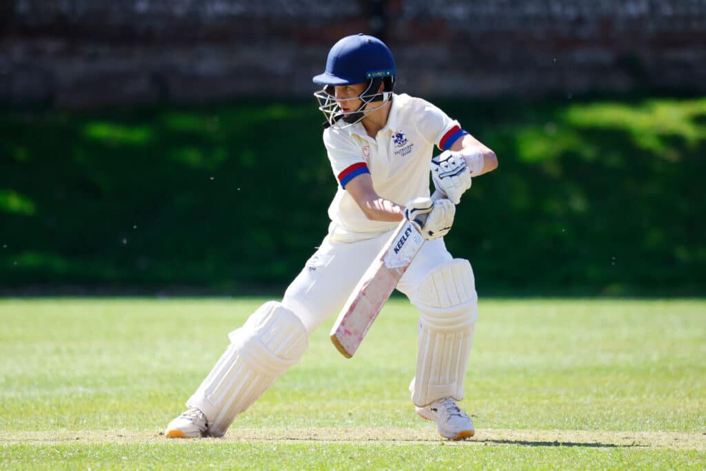 A cricket player in white kit and a blue helmet prepares to bat on a sunny pitch, focusing on the ball.