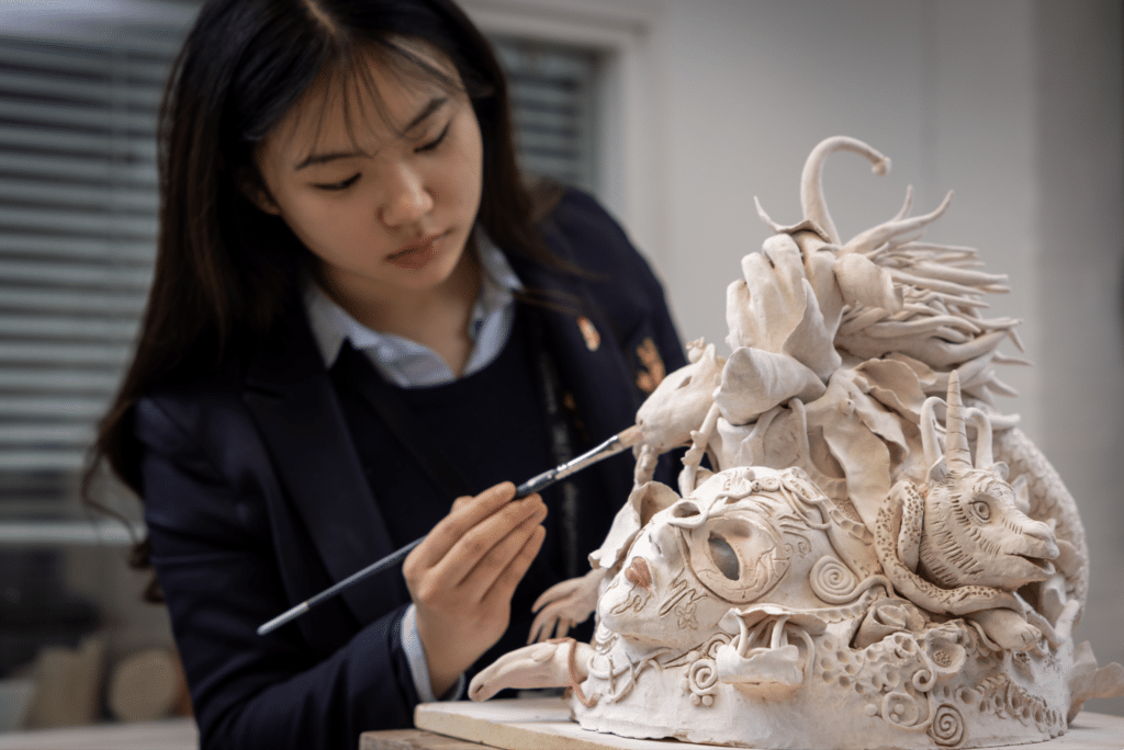 A young woman in a blazer carefully paints details on an intricate, fantasy-themed clay sculpture featuring animal and mythical creature faces.