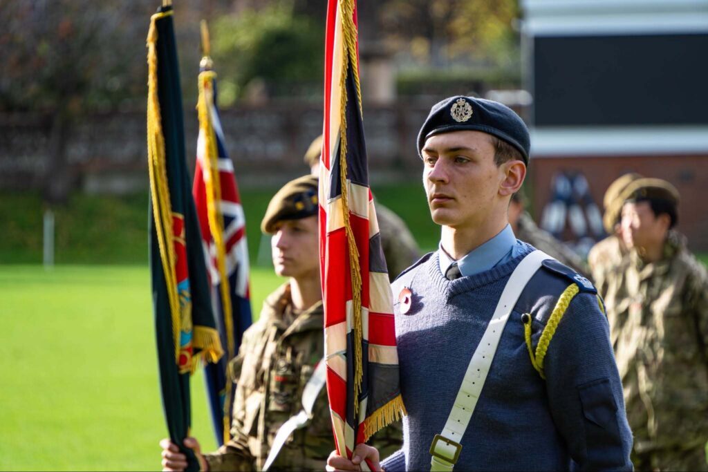 Young uniformed cadets stand at attention outdoors, holding flags during a ceremony on a sunny day, with one Eastbourne College cadet in focus wearing a blue beret and sash.