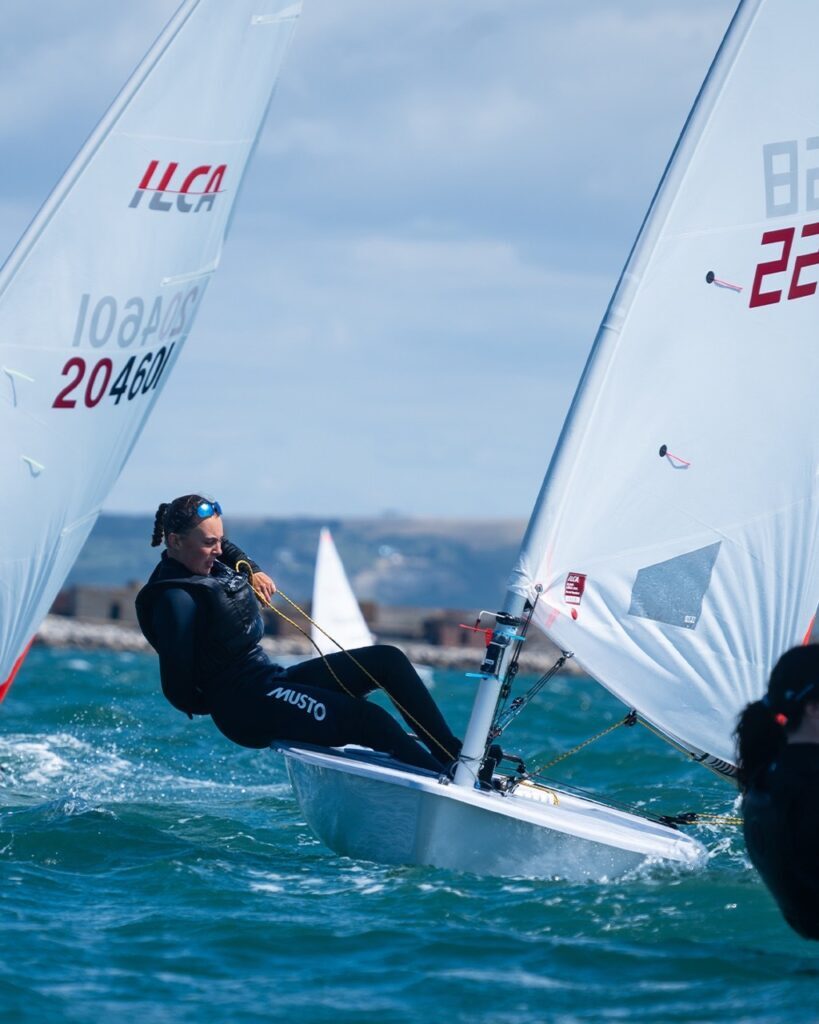 A person in a wetsuit sails a small ILCA dinghy on choppy blue water, leaning out to balance the boat as part of the British Sailing Pathway. Another sailboat and a distant shoreline are visible in the background under a cloudy sky.